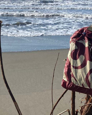 borsa a tracolla colore bordeaux appoggiata su ramo sulla spiaggia, mare sullo sfondo