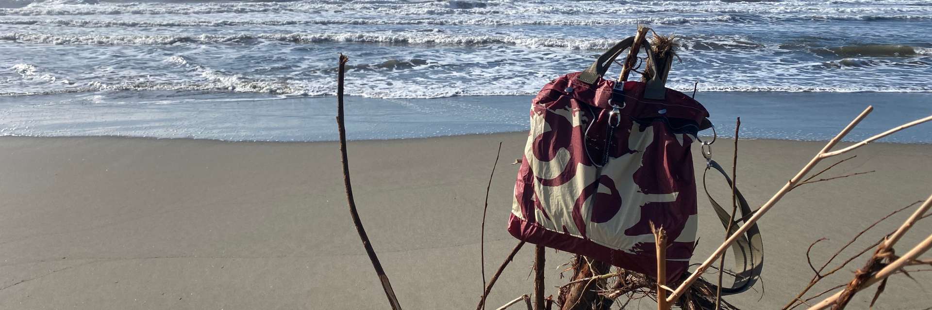 borsa a tracolla colore burgundy modello porta casco areonautica militare appoggiata su un ramo sulla spiaggia, mare sullo sfondo